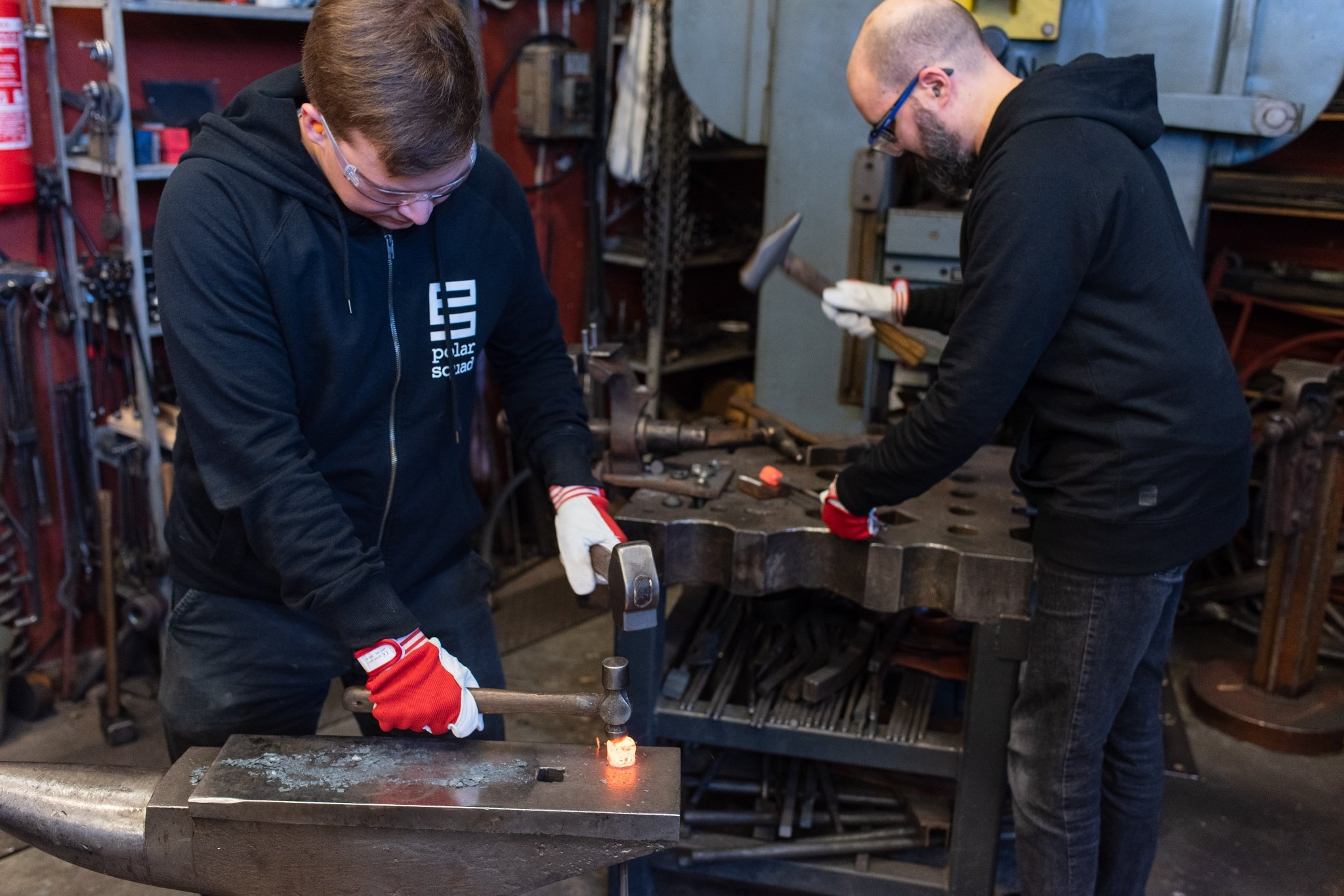 Two men hammering at billets, the one in front on an anvil, in the back on a table with templates